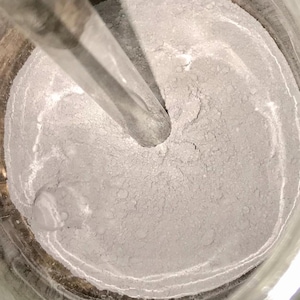 Op de afbeelding: Een close-up van een grijs poeder in een glazen container met een metalen roerstok. Het poeder wordt geroerd met de stok, waardoor een draaiend patroon ontstaat.