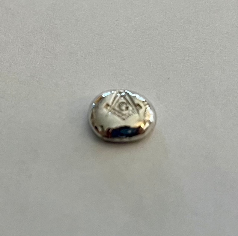 K&ouml;nnte beinhalten: Ein silberfarbener, ovaler Anh&auml;nger mit einem eingravierten Freimaurer-Symbol.