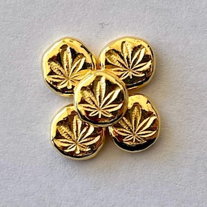 May include: Five gold-colored, round, cannabis leaf-shaped charms are arranged in a flower-like pattern. The charms have a shiny, reflective surface and are set against a light gray background. The cannabis leaf design is clearly visible.