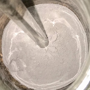 May include: A close-up of a metal container with a gray powder inside. A metal stirring rod is in the container, creating a swirl in the powder.