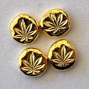 May include: Four gold-colored, round, leaf-shaped charms. Each charm has a detailed leaf design in the center. The charms are on a light gray surface.