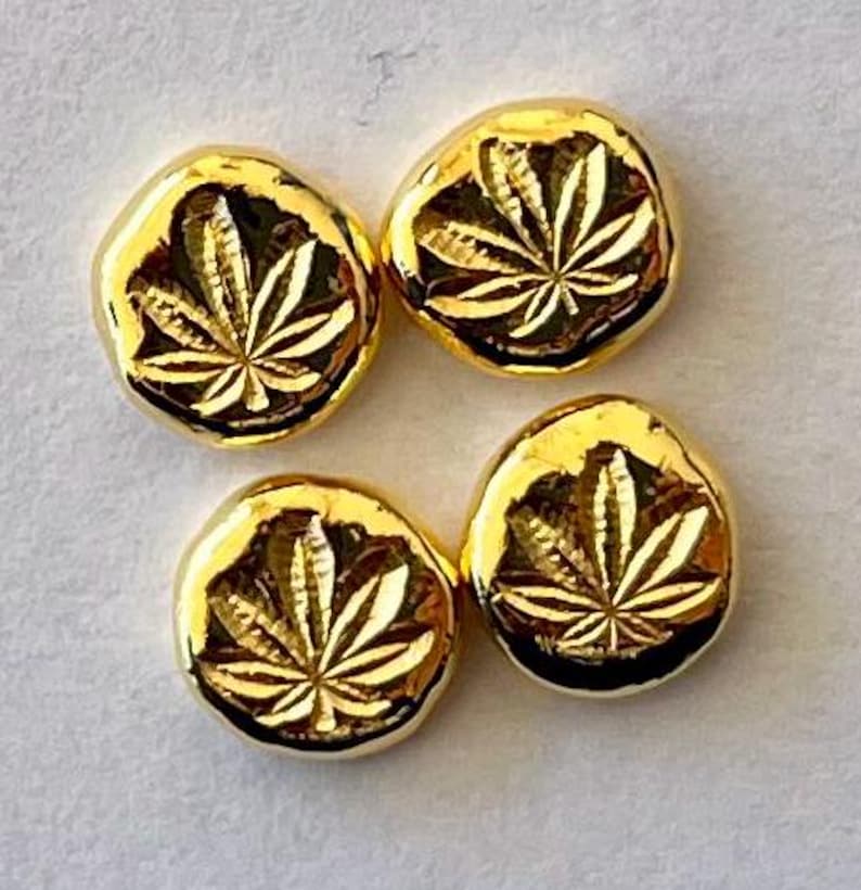 May include: Four gold-colored, round, leaf-shaped charms. Each charm has a detailed leaf design in the center. The charms are on a light gray surface.