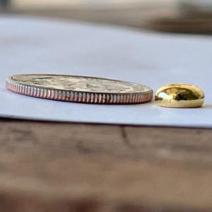 May include: A silver quarter coin with a ridged edge next to a small, smooth, gold-colored bead. The coin is flat and round, with a detailed design visible on its surface. The bead is a solid, rounded shape with a shiny finish.
