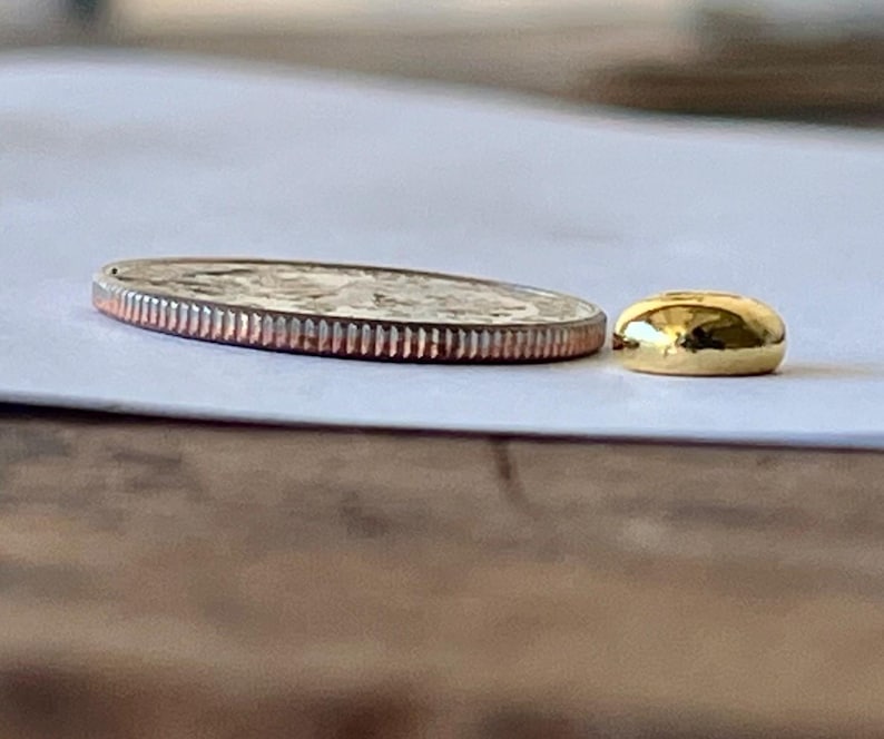 May include: A silver quarter coin with a ridged edge next to a small, smooth, gold-colored bead. The coin is flat and round, with a detailed design visible on its surface. The bead is a solid, rounded shape with a shiny finish.