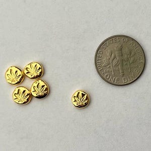May include: A collection of six small, gold-colored, round charms, each embossed with a cannabis leaf design. A US dime is included for size comparison. The charms are arranged on a white surface.