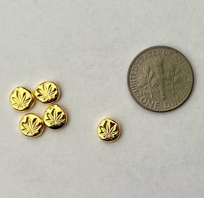 May include: A collection of six small, gold-colored, round charms, each embossed with a cannabis leaf design. A US dime is included for size comparison. The charms are arranged on a white surface.