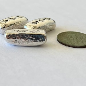 May include: Three silver bars, each with a textured surface and a stamped design, are displayed alongside a coin. The bars are marked with "10 GRAM" and a symbol. The coin is a dull green color.