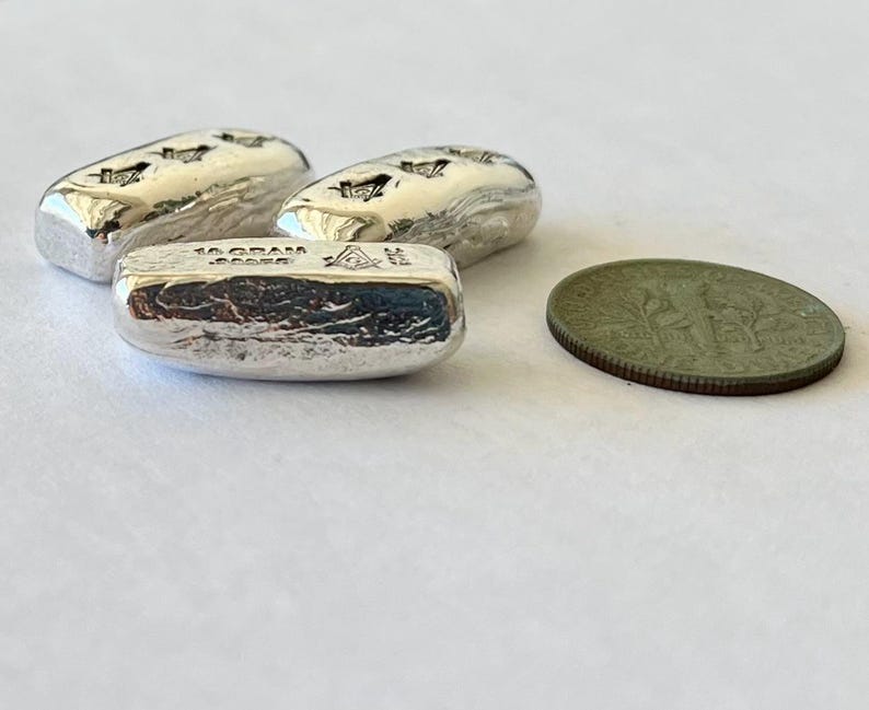May include: Three silver bars, each with a textured surface and a stamped design, are displayed alongside a coin. The bars are marked with "10 GRAM" and a symbol. The coin is a dull green color.