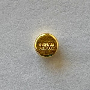 May include: A small, round, gold-colored object with the text "1 GRAM AU9999" stamped on its surface. The object has a slightly textured appearance and is set against a light-colored background. The gold color suggests a precious metal.