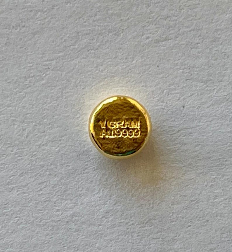 May include: A small, round, gold-colored object with the text "1 GRAM AU9999" stamped on its surface. The object has a slightly textured appearance and is set against a light-colored background. The gold color suggests a precious metal.