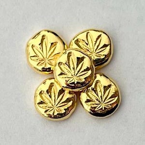 May include: Five gold-colored, round objects with a cannabis leaf design. The objects are arranged in a flower-like pattern, with each piece having a textured surface and a shiny finish. The background is a light, neutral color.