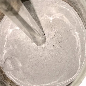 Puede incluir: Primer plano de un polvo gris en un recipiente de metal. Una varilla de agitación de metal está en el polvo, creando un patrón de remolino.