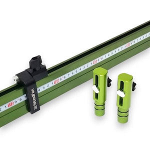 Op de afbeelding: Een groene WoodFan geleiderail voor een zaag met een liniaal aan de zijkant. De afbeelding toont ook twee groene verstelbare aanslagen. De geleiderail heeft een zwarte klem en het WoodFan-logo.