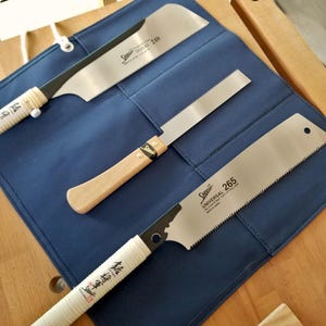 Può includere: Tre seghe giapponesi a trazione con manici in legno e bianchi sono esposte su un tessuto blu. Le seghe hanno lame argentate con il marchio "Shogun" e i numeri di modello stampati. Le seghe sono per la lavorazione del legno.