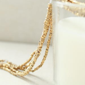 Può includere: Una collana composta da piccole perline color oro metallizzato con una texture sfaccettata. Il filo di perline è drappeggiato sul bordo di un contenitore di vetro trasparente, mostrando diversi giri di perline circolari uniformi e riflettenti.