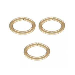 Pode incluir: Três argolas de metal em tom dourado, cada uma com formato oval e uma abertura visível no topo. Estes componentes de joalharia possuem uma superfície lisa e polida, dispostos em formação triangular sobre um fundo branco liso.