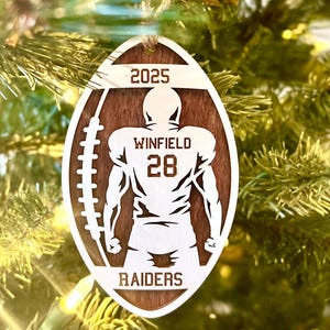 May include: A wooden, American football-shaped Christmas ornament. It features a football player silhouette, the name "WINFIELD", number "28", the word "RAIDERS", and the year "2025". The ornament is suspended from a Christmas tree.