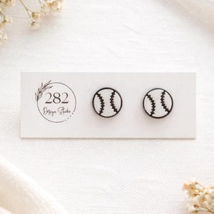 May include: A pair of baseball-themed stud earrings. Each earring features a black outline of a baseball with white stitching. The earrings are displayed on a white card with the text "282 Design Studio" and a floral design.