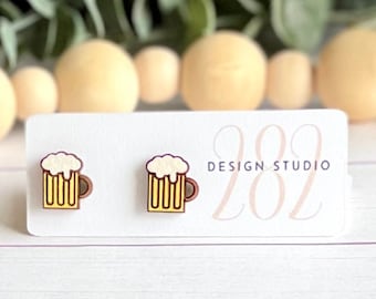 May include: A pair of wooden stud earrings featuring a beer mug design. The earrings have a yellow mug with a brown handle and a white foamy top. The earrings are on a white card with the text "202 Design Studio".