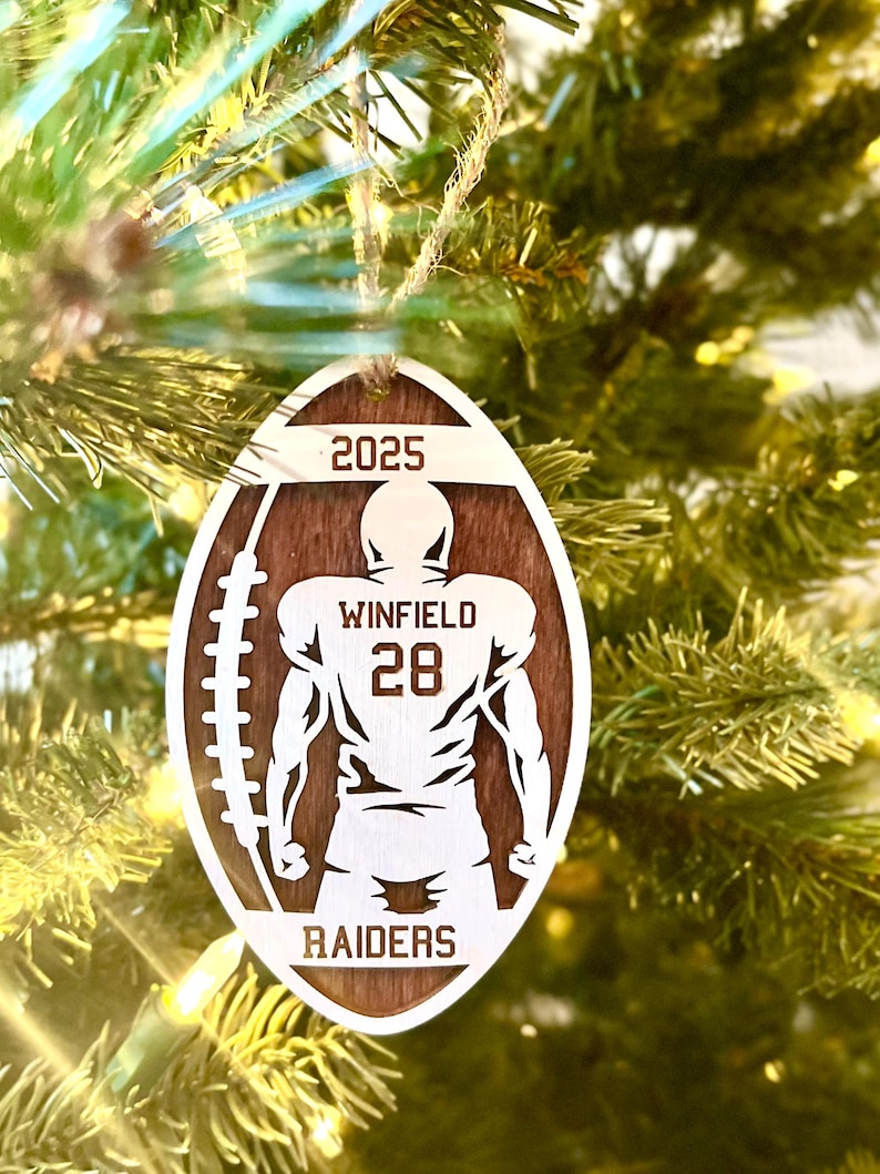 May include: A wooden, American football-shaped Christmas ornament. It features a football player silhouette, the name "WINFIELD", number "28", the word "RAIDERS", and the year "2025". The ornament is suspended from a Christmas tree.