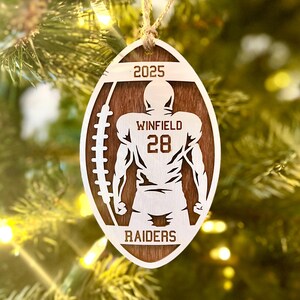 May include: A wooden football-shaped Christmas ornament featuring a football player silhouette. The ornament displays the text "2025", "WINFIELD 28", and "RAIDERS". It is suspended by a twine string, set against a blurred Christmas tree backdrop.