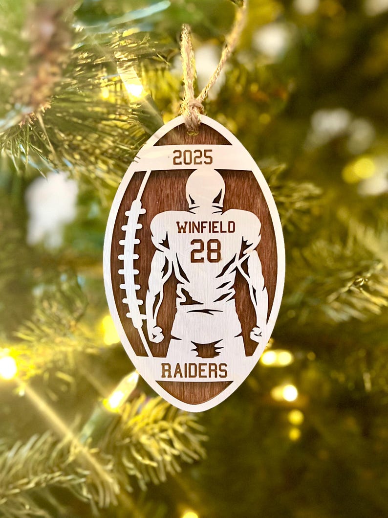 May include: A wooden football-shaped Christmas ornament featuring a football player silhouette. The ornament displays the text "2025", "WINFIELD 28", and "RAIDERS". It is suspended by a twine string, set against a blurred Christmas tree backdrop.