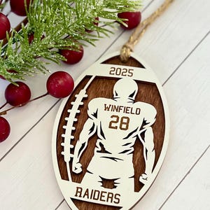 May include: Wooden football-shaped ornament featuring a football player silhouette. The ornament is brown with white detailing, including the text "2025", "Winfield 28", and "Raiders". A twine loop is attached for hanging.
