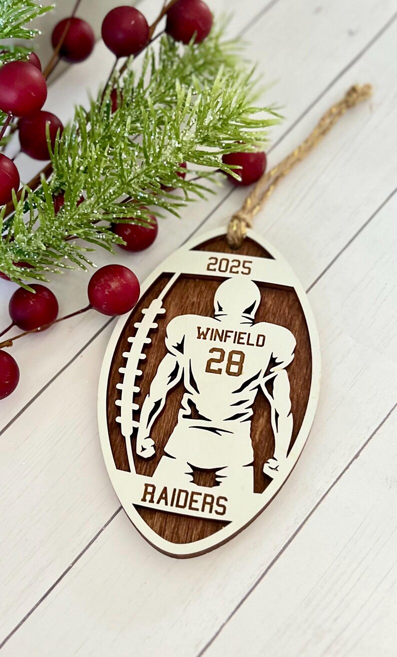 May include: Wooden football-shaped ornament featuring a football player silhouette. The ornament is brown with white detailing, including the text "2025", "Winfield 28", and "Raiders". A twine loop is attached for hanging.