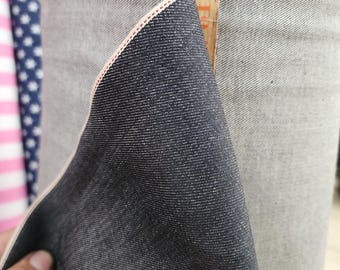 May include: Close-up of a roll of dark indigo denim fabric. The fabric is folded over, revealing a pink edge. A wooden ruler with inch markings is placed next to the fabric. The denim has a textured appearance.