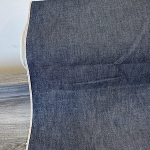May include: A rolled-up bolt of dark blue denim fabric. The fabric has a textured appearance and a visible selvedge edge with a light-colored stripe. The denim is folded over, revealing the fabric's thickness and texture.