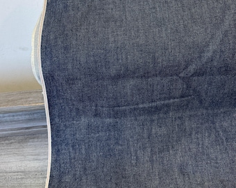 May include: A rolled-up bolt of dark blue denim fabric. The fabric has a textured appearance and a visible selvedge edge with a light-colored stripe. The denim is folded over, revealing the fabric's thickness and texture.