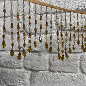 May include: Decorative beaded curtain with clear, amber, and gold beads. The beads are strung on gold-colored wire and hang from a curved, gold-colored wooden frame. The background is a white brick wall.