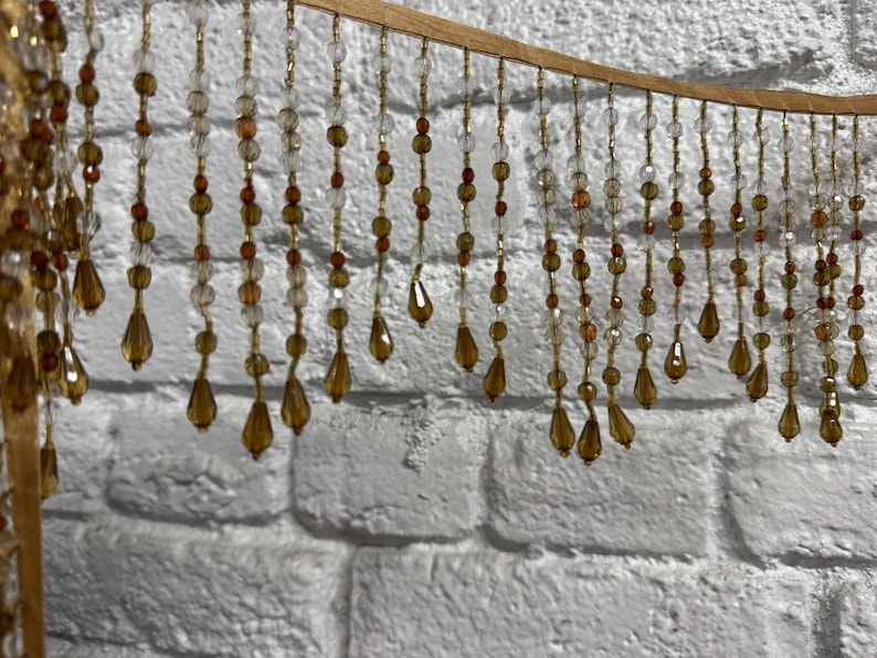 May include: Decorative beaded curtain with clear, amber, and gold beads. The beads are strung on gold-colored wire and hang from a curved, gold-colored wooden frame. The background is a white brick wall.
