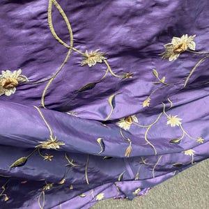 May include: Close-up of a purple fabric with embroidered floral design. The embroidery features gold-colored stems, leaves, and flowers with cream-colored petals. The fabric has a shiny, silky texture and is folded in layers.