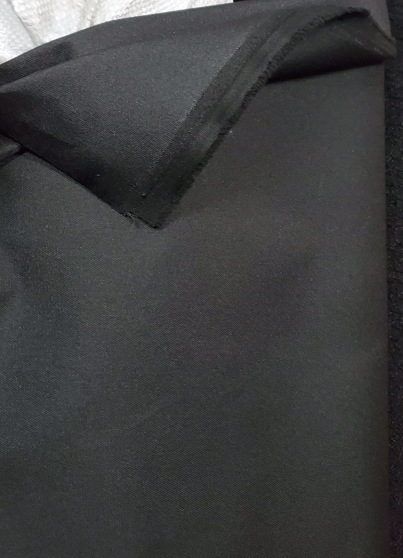 May include: Black fabric with a smooth, slightly textured surface. The fabric is folded over, showing the raw edge.
