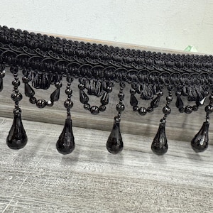 Può includere: Bordura di frange nere con perline con pendenti a forma di goccia. La bordura presenta una base intrecciata con dettagli ad anello e piccole perline, e i pendenti sono appesi sotto. Questa bordura decorativa è adatta a vari progetti artigianali.