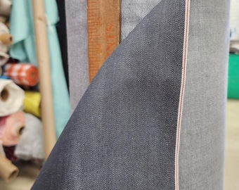 May include: A roll of dark gray denim fabric with a red selvedge edge. A wooden ruler is placed vertically in the center of the roll. The fabric is tightly rolled, showcasing the texture and color variations of the denim.