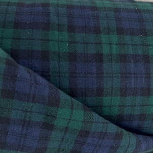 Puede incluir: Un rollo de tela con un patrón de cuadros escoceses clásico en tonos profundos de verde bosque, azul marino y negro. El tejido tiene una textura visible con una esquina doblada que revela el reverso del material y bordes deshilachados.
