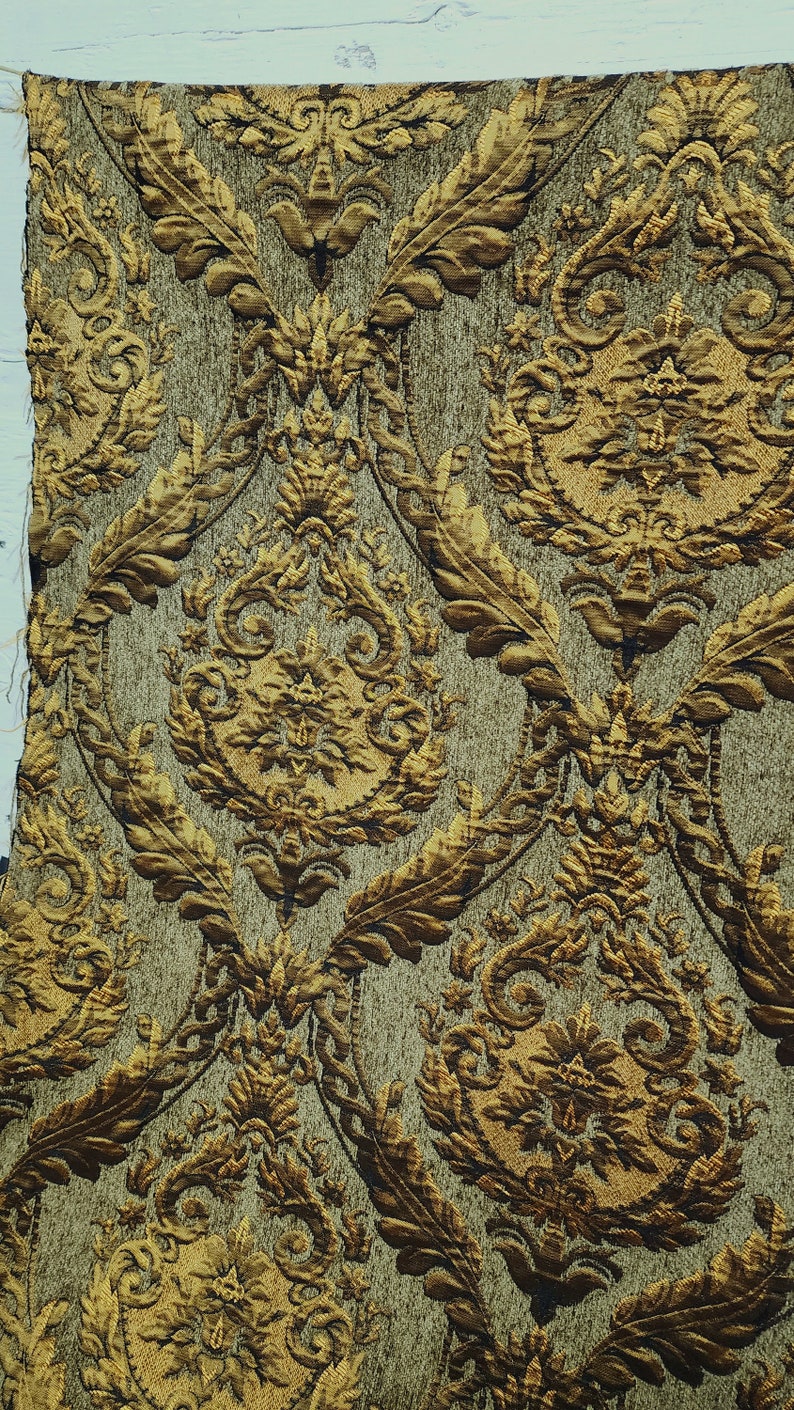 May include: A close-up of a fabric with a gold and brown damask pattern. The fabric has a textured surface and a repeating pattern of leaves and flowers.