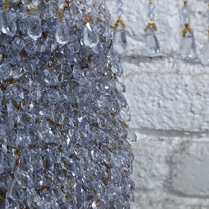 Op de afbeelding: Close-up van een decoratief kroonluchterelement. Het stuk is bedekt met heldere, facetgeslepen kristallen, met goudkleurige accenten. Hangende strengen kristallen zijn zichtbaar tegen een witte bakstenen muur, wat een sprankelend, elegant effect creëert.