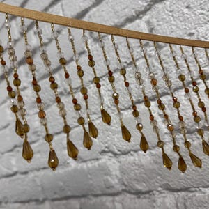 May include: A decorative beaded fringe with a wooden top. The fringe features strands of clear, amber, and gold beads, including teardrop-shaped pendants. The beads are strung on gold-colored wire, creating a shimmering effect.