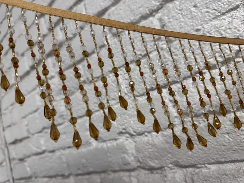 May include: A decorative beaded fringe with a wooden top. The fringe features strands of clear, amber, and gold beads, including teardrop-shaped pendants. The beads are strung on gold-colored wire, creating a shimmering effect.