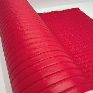 Op de afbeelding: Een rol felrode gewatteerde stof met horizontale stiksels. De stof is gedeeltelijk uitgerold en toont een witte, donzige achterkant. Het materiaal heeft een glanzende afwerking en lijkt van gemiddeld gewicht te zijn, geschikt voor diverse naaiprojecten.