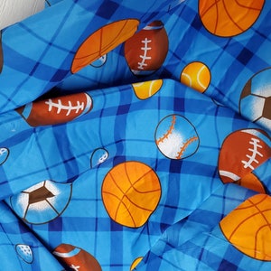 May include: A blue fabric featuring a plaid pattern overlaid with a repeating print of various sports balls including orange basketballs, brown footballs, white soccer balls, yellow tennis balls, and white baseballs with orange stitching.