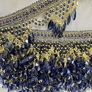 May include: Navy blue and gold beaded decorative trim. The trim has a woven base with rows of small beads and dangling, faceted, navy blue and gold crystal-like beads. Suitable for embellishing clothing or home decor. The trim is approximately 10 cm wide.