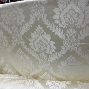 May include: Ivory-colored fabric with a detailed damask pattern. The fabric features a repeating design of floral and scroll motifs in a lighter shade, creating a subtle contrast. The material appears to be draped, showcasing its texture and flow.