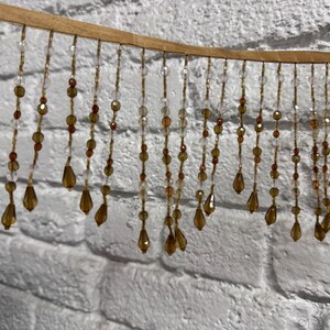 May include: A decorative beaded fringe with a tan backing. The fringe features strands of clear, amber, and brown beads, including teardrop-shaped pendants. The beads hang in a vertical pattern against a white brick wall.