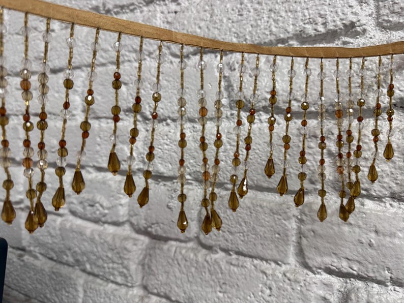 May include: A decorative beaded fringe with a tan backing. The fringe features strands of clear, amber, and brown beads, including teardrop-shaped pendants. The beads hang in a vertical pattern against a white brick wall.