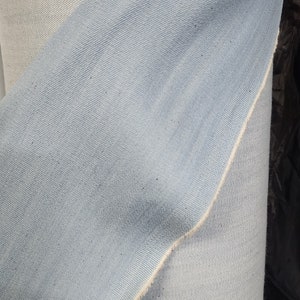 May include: Close-up of a light blue denim fabric with a white seam. The fabric is folded over itself, showing the texture of the denim.