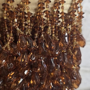 May include: Close-up of decorative trim featuring strands of faceted, amber-colored beads. The beads hang from a gold-colored fabric backing. The beads are teardrop-shaped and reflect light.